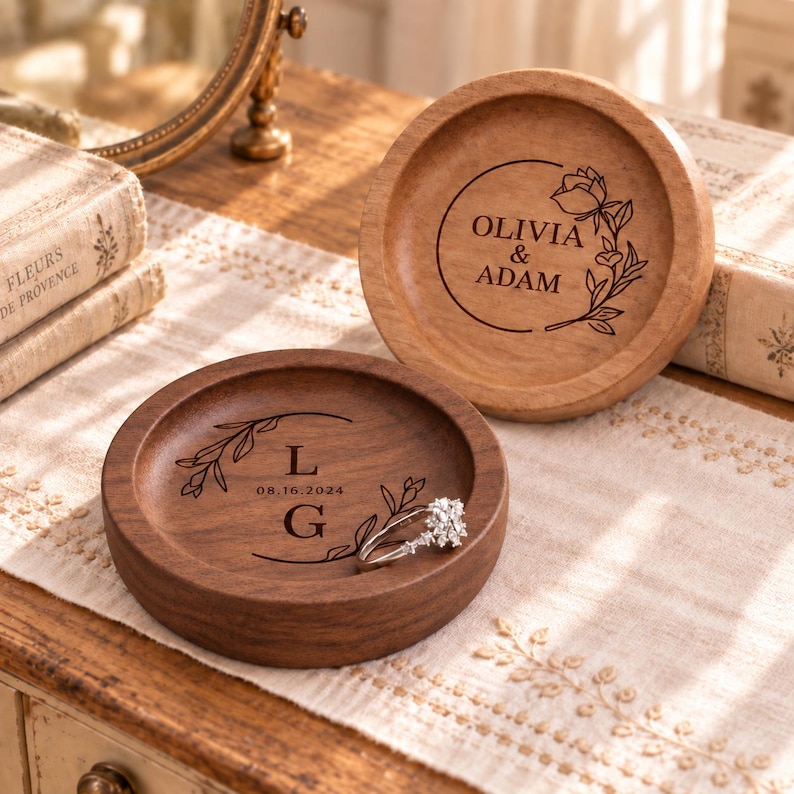 May include: Two round wooden ring dishes with engraved designs. The lighter dish features a floral circle with the text OLIVIA & ADAM. The darker walnut dish displays the initials L and G with the date 08.16.2024 and a silver ring inside.