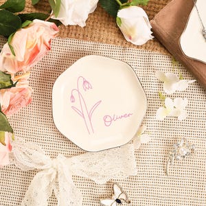 May include: A cream-colored, irregularly shaped ceramic plate with a black rim. The plate features a lavender floral design and the name "Oliver" in a matching script. The plate is surrounded by flowers and decorative elements.