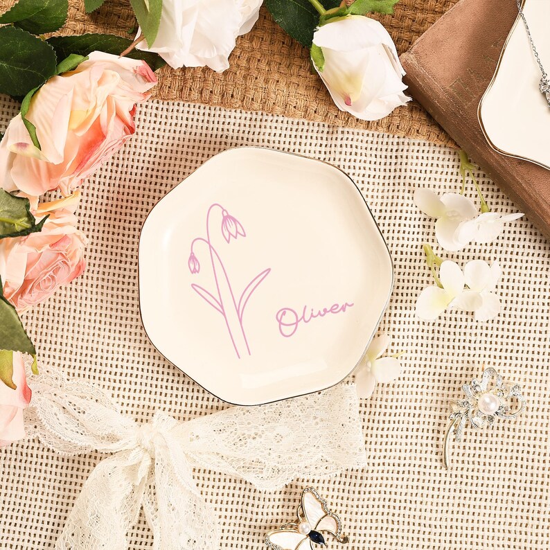 May include: A cream-colored, irregularly shaped ceramic plate with a black rim. The plate features a lavender floral design and the name "Oliver" in a matching script. The plate is surrounded by flowers and decorative elements.