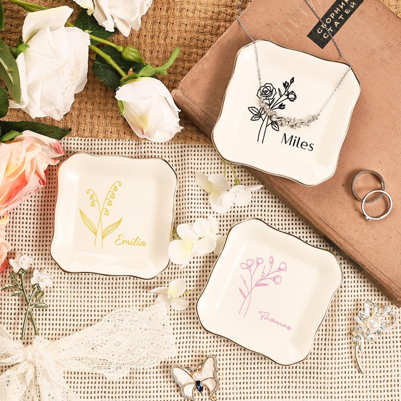 May include: Three off-white ceramic jewelry dishes with gold trim. Each dish features a floral design and a name: "Miles", "Emilia", and "Thomas". A silver necklace, rings, and a decorative butterfly are also present.