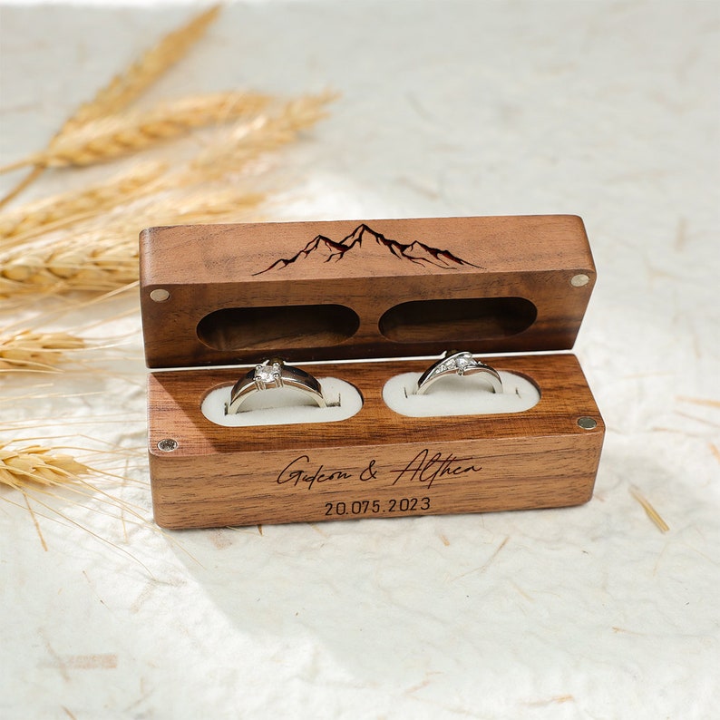 May include: A wooden ring box with a mountain design engraved on the lid. The box has two compartments for rings and is personalised with the names "Gysen & Althen" and the date "20.07.5.2023".