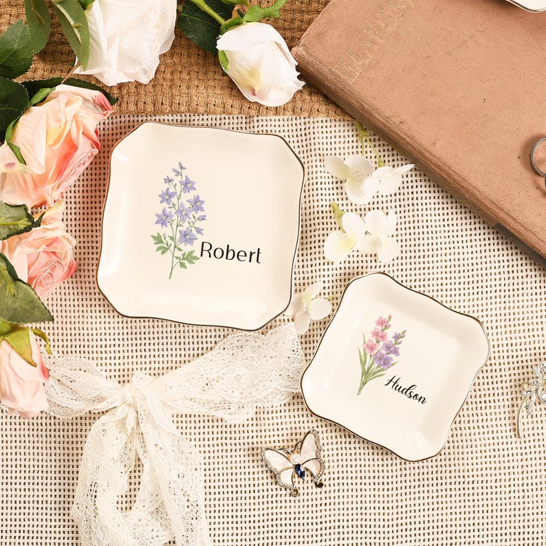May include: Two off-white square trinket dishes with a floral design and black trim. One dish has the name "Robert" and the other has the name "Hudson". The dishes are surrounded by flowers and a decorative butterfly pin.