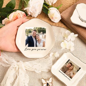 Puede incluir: Plato de cer&aacute;mica blanquecina con borde dorado, con una foto de una pareja y el texto "Love you forever". El plato est&aacute; sostenido por una mano, rodeado de flores y otros objetos decorativos. Se ve un segundo plato.
