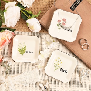 May include: Three square, off-white ceramic trinket dishes with gold trim. Each dish features a floral design and a name: "Charles," "Victoria," and "Stella." A necklace and rings are on the dishes. The background includes flowers and a book.