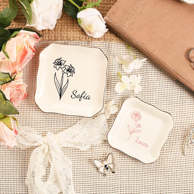May include: Two off-white square trinket dishes with floral designs and names. One dish has a black floral design with the name "Sofia", and the other has a pink floral design with the name "Lucas". Both dishes have a gold trim.