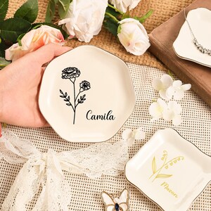 May include: Off-white ceramic trinket dish with a gold trim, featuring a black floral design and the name "Camila". The dish has an irregular, hexagonal shape. Other similar dishes are also visible.