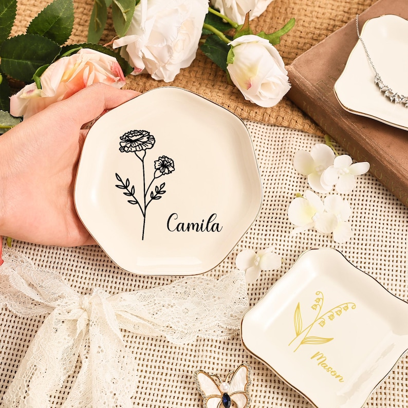 May include: Off-white ceramic trinket dish with a gold trim, featuring a black floral design and the name "Camila". The dish has an irregular, hexagonal shape. Other similar dishes are also visible.