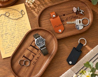 Op de afbeelding: Twee ovale houten schalen met gegraveerde tekst. Eén schaal toont "Julia & Peter" met een beige leren sleutelhanger, zilveren manchetknopen en een sleutelring. De andere toont "OLIV 10.16.20" met een polshorloge en twee zilveren ringen.