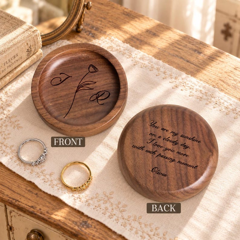 May include: A round wooden ring dish with a dark brown finish. The interior features an engraved flower and the letters J and R. The exterior base is engraved with the text: You are my sunshine on a cloudy day. I love you more with each passing moment. Olivia.