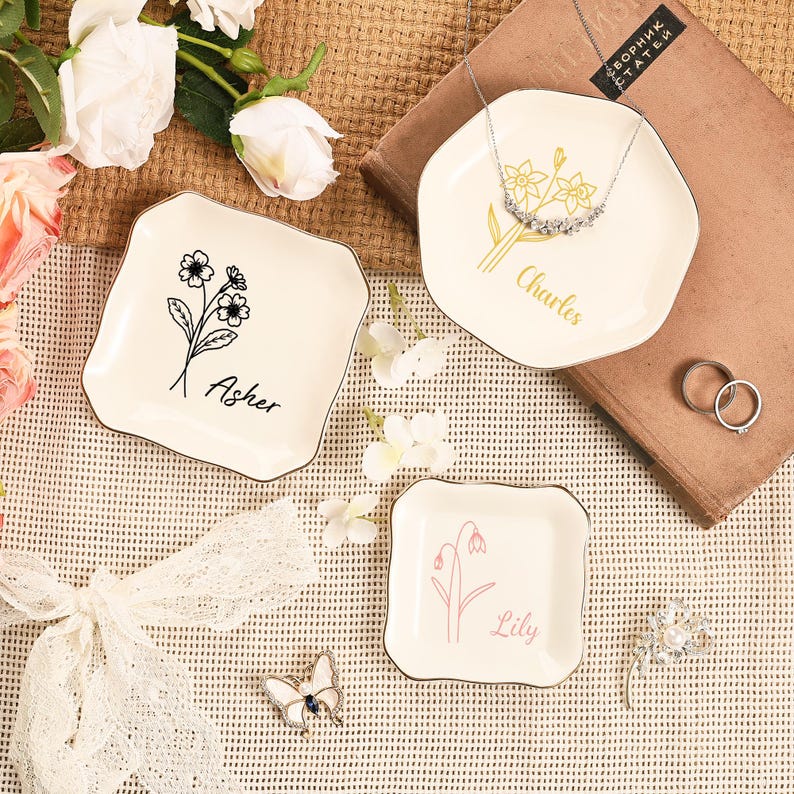 May include: Three off-white decorative trays with floral designs and names. One tray has a black floral design with the name "Asher". Another has a gold floral design with the name "Charles". The third has a pink floral design with the name "Lily".