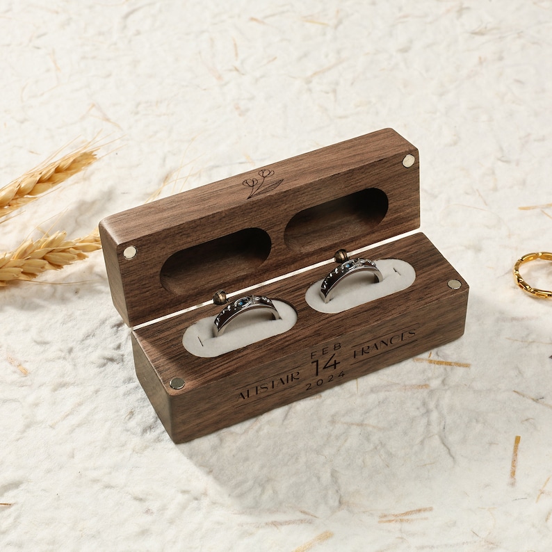 May include: A wooden ring box with two compartments for rings. The box is engraved with the names "Alistair" and "Frances", the date "Feb 14, 2024", and a small floral design.