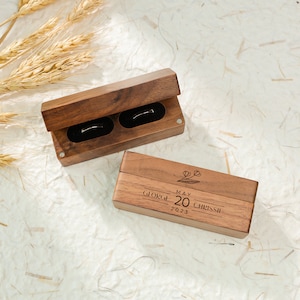 May include: Two wooden ring boxes. The top box is open and shows two empty ring slots. The bottom box is closed and has the text "GEORGE MAY 20 CHRISSI 2023" engraved on the top. The boxes are made of dark wood and have a natural finish.
