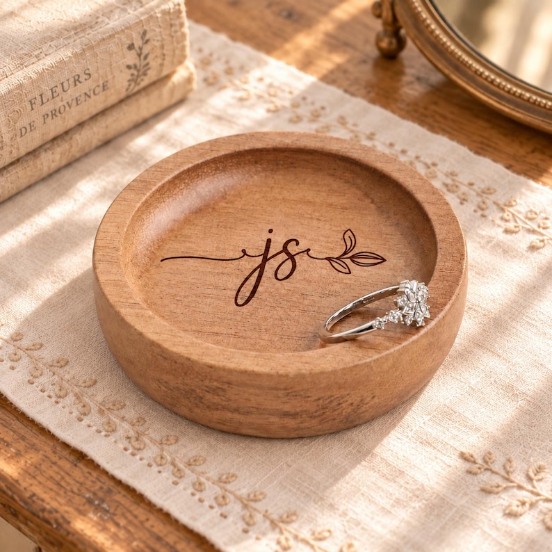 May include: A round wooden trinket dish with a smooth, light brown finish. The center of the dish features a dark brown laser-engraved monogram of the letters js connected by a decorative flourish and a small leaf design. A silver ring rests inside.