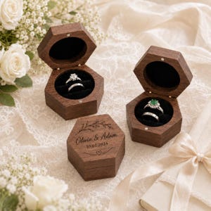 Op de afbeelding: Drie zeshoekige ringdoosjes van donker hout met een zwarte fluwelen binnenkant. Twee doosjes staan open en bevatten elk twee zilveren ringen. Het derde doosje is gesloten en gegraveerd met de tekst Olivia & Adam, de datum 10.02.2024 en een bladmotief.