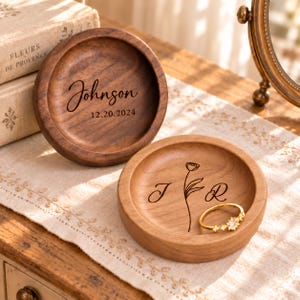 May include: Two round wooden trinket dishes with engraved details. One dish features the name Johnson and the date 12.20.2024. The other dish displays the initials J and R with a flower illustration and contains a gold ring with small stones.
