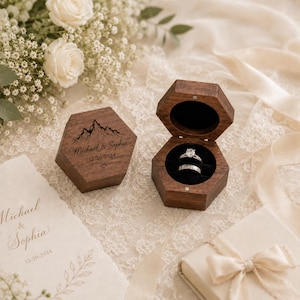 May include: Two hexagonal dark wood ring boxes. One box is closed, featuring an engraved mountain graphic, the text Michael & Sophia, the date 12.20.2024, and a heart. The other box is open, revealing two silver rings inside a black velvet insert.