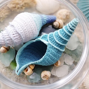 Peut inclure: Deux décorations de coquillages crochetés dans un bocal en verre. L'une est blanche et bleue, l'autre bleu clair. Le bocal contient du sable, de petits coquillages et du verre de mer. Thème côtier.
