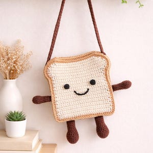Può includere: Una borsa all'uncinetto fatta a mano a forma di fetta di pane tostato. La borsa presenta un volto sorridente, braccia e gambe marroni, con un cinturino abbinato. Un accessorio unico e divertente.