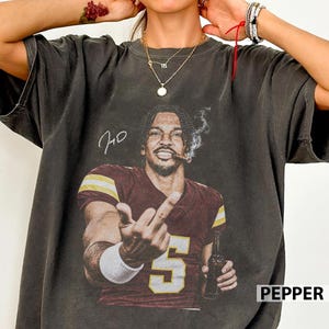 May include: A charcoal grey short-sleeve t-shirt featuring a graphic print of a football player in a burgundy and gold jersey with the number 5. The player is holding a bottle and gesturing with a middle finger. The text "JMD" is printed above.