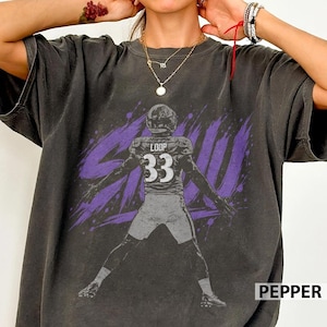 May include: A charcoal grey short-sleeve t-shirt featuring a graphic of a football player wearing jersey number 33 with the name LOOP on the back. The design includes a purple brushstroke background and a distressed, vintage-inspired aesthetic.