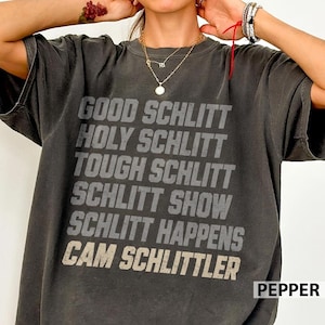 May include: A charcoal grey short-sleeved t-shirt featuring distressed light grey text that reads: GOOD SCHLITT, HOLY SCHLITT, TOUGH SCHLITT, SCHLITT SHOW, SCHLITT HAPPENS, CAM SCHLITTLER. The shirt has a crew neckline and a relaxed, vintage-inspired fit.