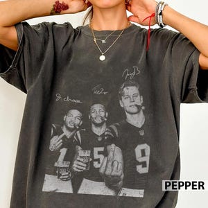 May include: A charcoal gray short-sleeve t-shirt featuring a vintage-style graphic print of three football players in jerseys with the numbers 1, 5, and 9. The design includes printed signatures and the players are shown holding drinks.