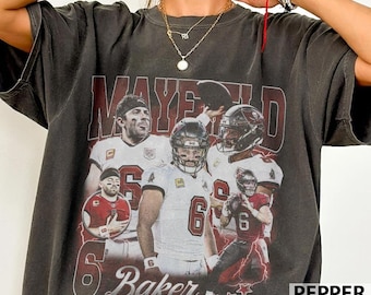 Puede incluir: Una camiseta de manga corta gris carbón con un estampado gráfico de un jugador de fútbol americano en varias poses. El texto "MAYFIELD" está impreso en letras rojas grandes en la parte superior, con el número "6" y "Baker" debajo.