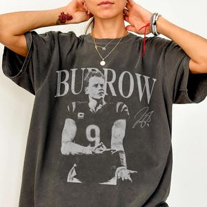 May include: A charcoal grey short-sleeve t-shirt featuring a large graphic print of a football player in a jersey with the number 9. The text BURROW is printed in large capital letters above the player, accompanied by a signature graphic.