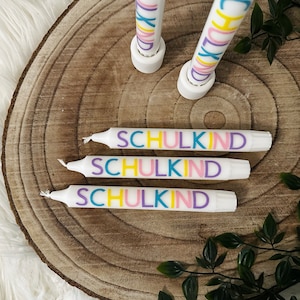 May include: White candles with the word "SCHULKIND" in colorful letters. The candles are arranged on a wooden surface with a natural wood grain pattern. Green leaves are in the background.