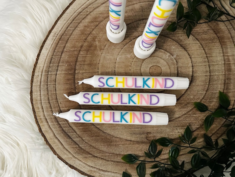 May include: White candles with the word "SCHULKIND" in colorful letters. The candles are arranged on a wooden surface with a natural wood grain pattern. Green leaves are in the background.
