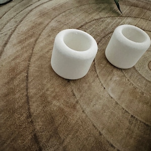 May include: Two small, white, cylindrical candle holders. The holders are made of a textured material and have a hollowed-out center. They are placed on a light brown, wood-grain surface, with green foliage in the background.