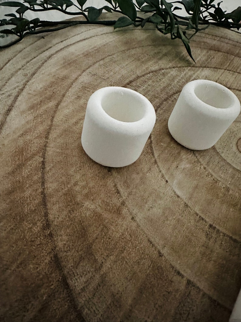 May include: Two small, white, cylindrical candle holders. The holders are made of a textured material and have a hollowed-out center. They are placed on a light brown, wood-grain surface, with green foliage in the background.