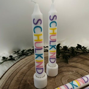 May include: White candles with the word "SCHULKIND" printed in colorful letters. The candles are standing on white holders, with a wooden background. The word "SCHULKIND" is printed in purple, yellow, pink, and blue.