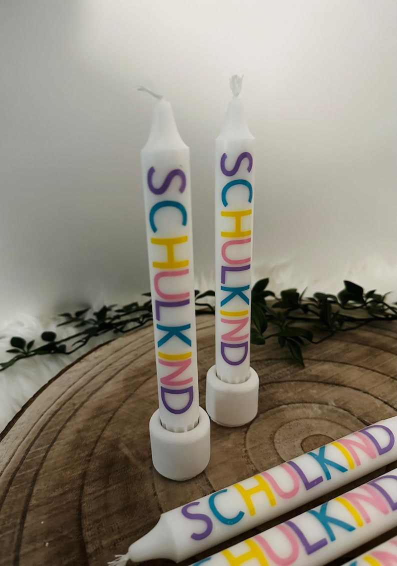 May include: White candles with the word "SCHULKIND" printed in colorful letters. The candles are standing on white holders, with a wooden background. The word "SCHULKIND" is printed in purple, yellow, pink, and blue.