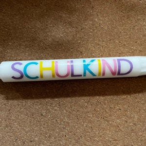 May include: White candle with the word "SCHULKIND" in colorful letters. The candle has a wick at one end and is resting on a brown surface. The text is in purple, yellow, pink, and blue.