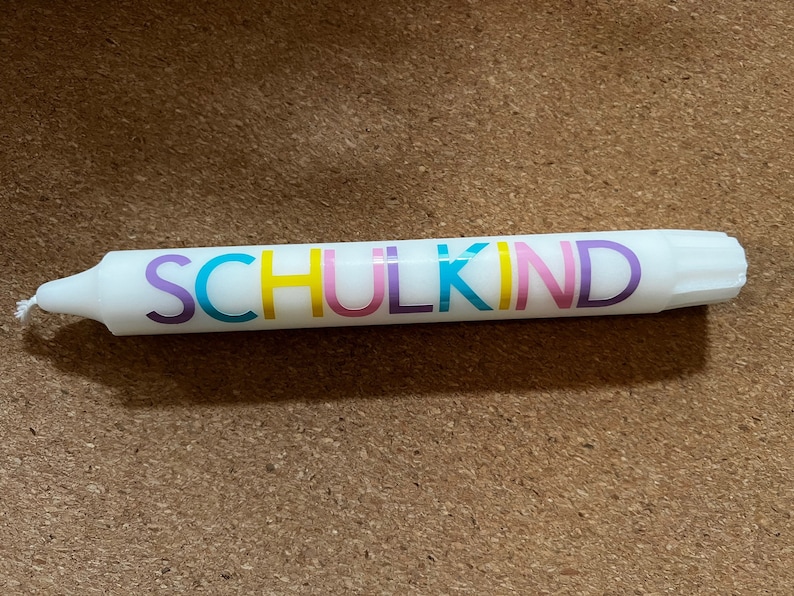 May include: White candle with the word "SCHULKIND" in colorful letters. The candle has a wick at one end and is resting on a brown surface. The text is in purple, yellow, pink, and blue.