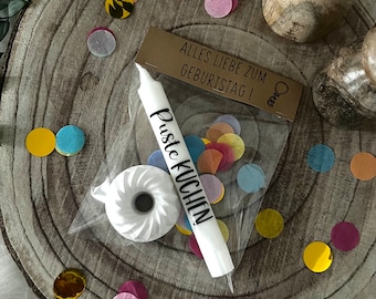 May include: A birthday-themed gift set on a wooden slice, featuring a white candle with "Puste Kuchen" text, a small white cake, confetti, and a card with German text. The confetti is in various colors.