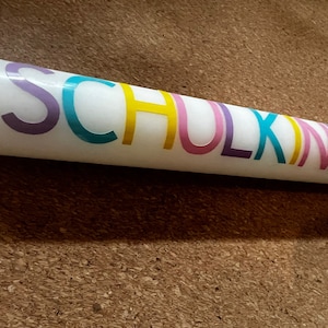 May include: A white candle with the word "SCHULKIND" printed in colorful letters. The candle is cylindrical and appears to be made of wax. The text is in a rainbow of colors, including purple, teal, yellow, pink, and purple.