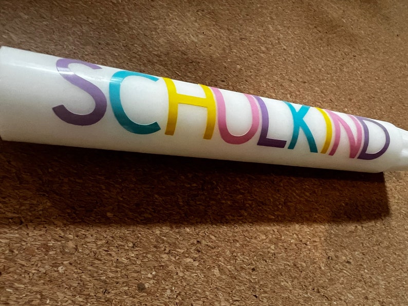 May include: A white candle with the word "SCHULKIND" printed in colorful letters. The candle is cylindrical and appears to be made of wax. The text is in a rainbow of colors, including purple, teal, yellow, pink, and purple.