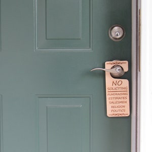 May include: A wooden door hanger with the words "NO SOLICITING" and other categories printed on it, hanging on a teal door. The door has a silver handle and a lock.