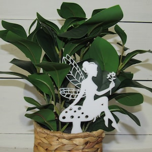 May include: White silhouette of a fairy sitting on a mushroom with wings spread out. The fairy is holding a flower in her hand. The fairy is in front of a potted green plant.