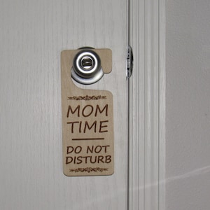 May include: A wooden door hanger with the text "MOM TIME DO NOT DISTURB" in brown lettering. The hanger has a silver hook and a decorative design.