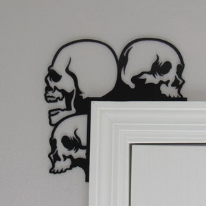 May include: Three black metal skull silhouettes are mounted on a white door frame. The skulls are arranged in a corner, with two skulls facing forward and one skull facing sideways.