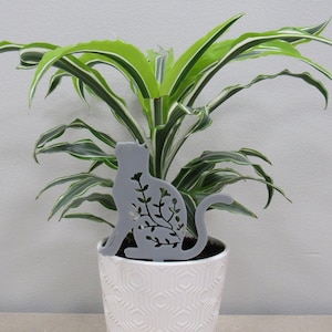 May include: A gray metal cat silhouette with floral cutouts, placed in a white ceramic pot with a green and white plant. The cat is sitting upright, with its tail curled. The plant has long, pointed leaves.