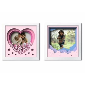 Peut inclure: Deux cadres photo blancs avec un design en papier d&eacute;coup&eacute; en 3D. Le cadre de gauche a un c&oelig;ur rose avec une photo de famille &agrave; l'int&eacute;rieur. Le cadre de droite a un design de nuage bleu et rose avec une photo d'un enfant courant dans un champ.