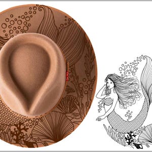 May include: A tan fedora hat with a mermaid design etched on the brim. The hat features a light brown crown and a detailed illustration of a mermaid with flowing hair and a fish tail. The brim also has an underwater scene.