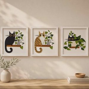 May include: A set of three square white-framed wall art pieces featuring layered cutouts of cats and green ivy plants. The first shows a black cat, the second a brown tabby cat, and the third a black cat peeking through a dense cluster of green leaves.