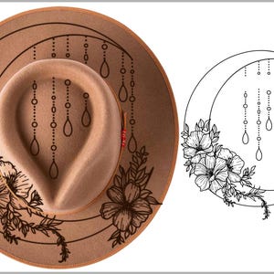 May include: A tan fedora hat with a floral design and a crescent moon illustration. The hat features a detailed black floral pattern on the brim and crown. The crescent moon design includes flowers and hanging teardrop shapes.