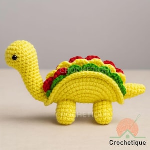Puede incluir: Una tortuga de ganchillo amarilla con un cuerpo de concha de taco. La concha del taco está rellena de hilo rojo, verde y amarillo, que se asemeja a los rellenos del taco. La tortuga tiene un cuello largo, patas pequeñas y una cola. La palabra "Crochetique" está en la esquina inferior derecha.