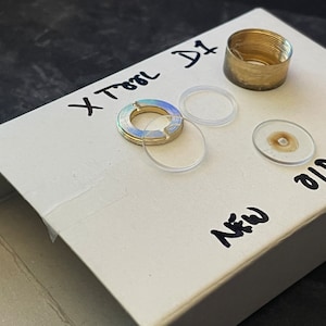 以下が含まれることがあります： 「X TOOL D1」と「NEW OLD」と書かれた白い箱。箱の中には、金色の金属片、透明なプラスチック製のリングが2つ、中央に茶色の斑点がある透明なプラスチック製の円盤が入っています。